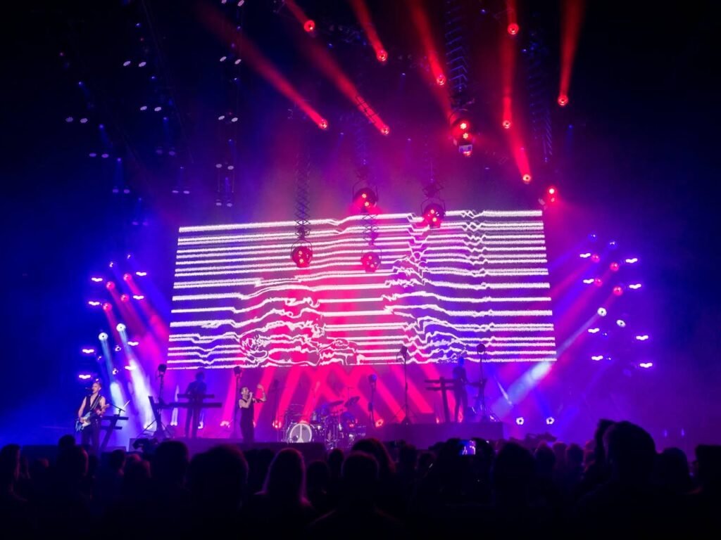 A live concert performance with vivid pink and purple stage lighting and a large animated LED screen.