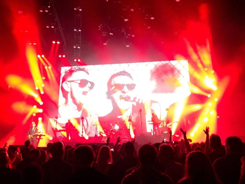 Live concert performance with vibrant red lighting and a large stage screen. Silhouetted crowd watching and raising hands.
