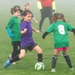 Young players in green and purple jerseys compete for a soccer ball during a foggy match.