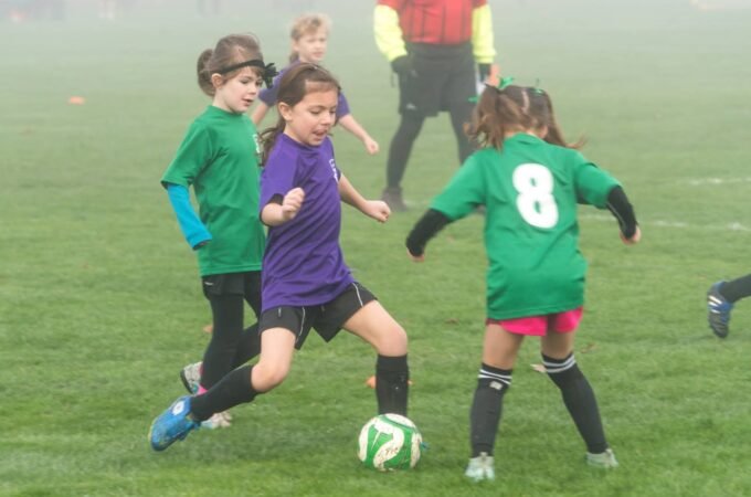 Young players in green and purple jerseys compete for a soccer ball during a foggy match.