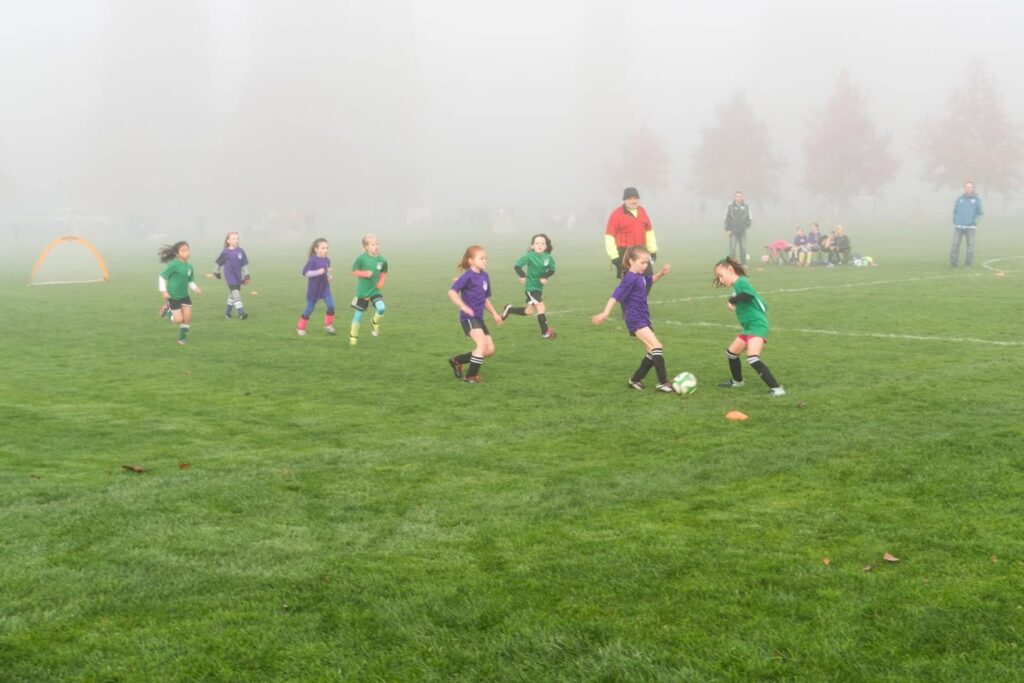 Two images showing a group of children playing a game of soccer on a large gravel field during a morning fog.