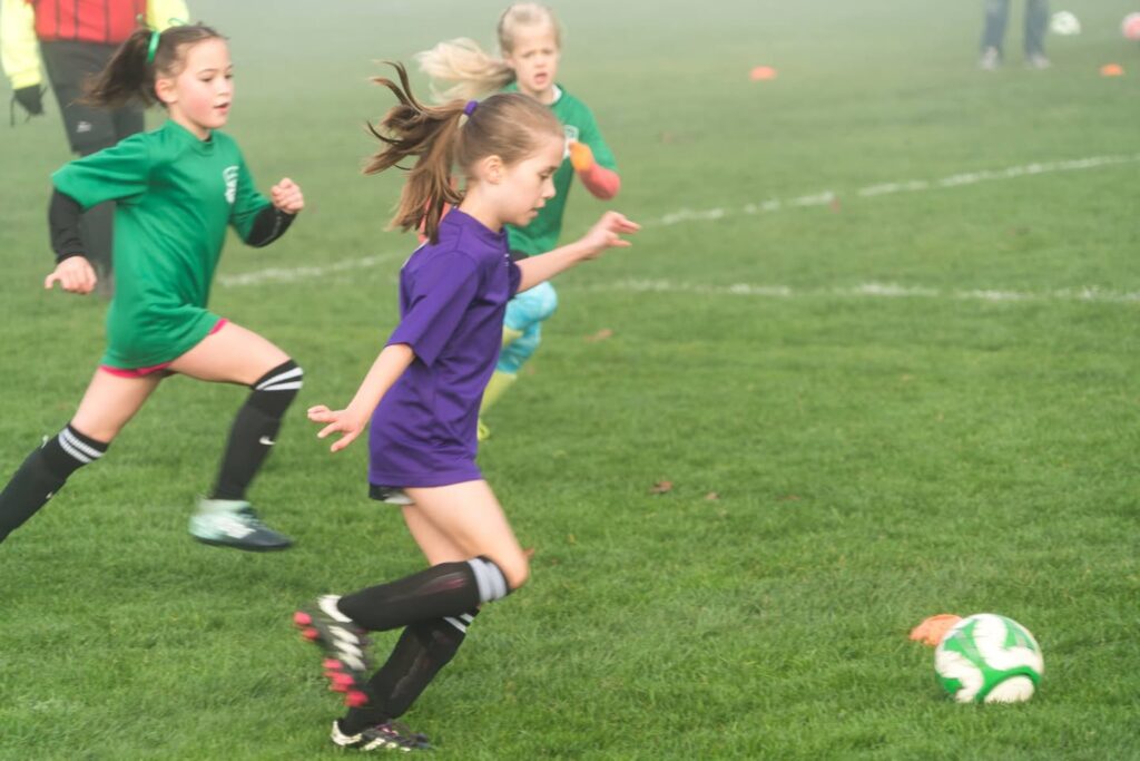 A dynamic action shot of youth soccer players on a grass field in green and purple uniforms.