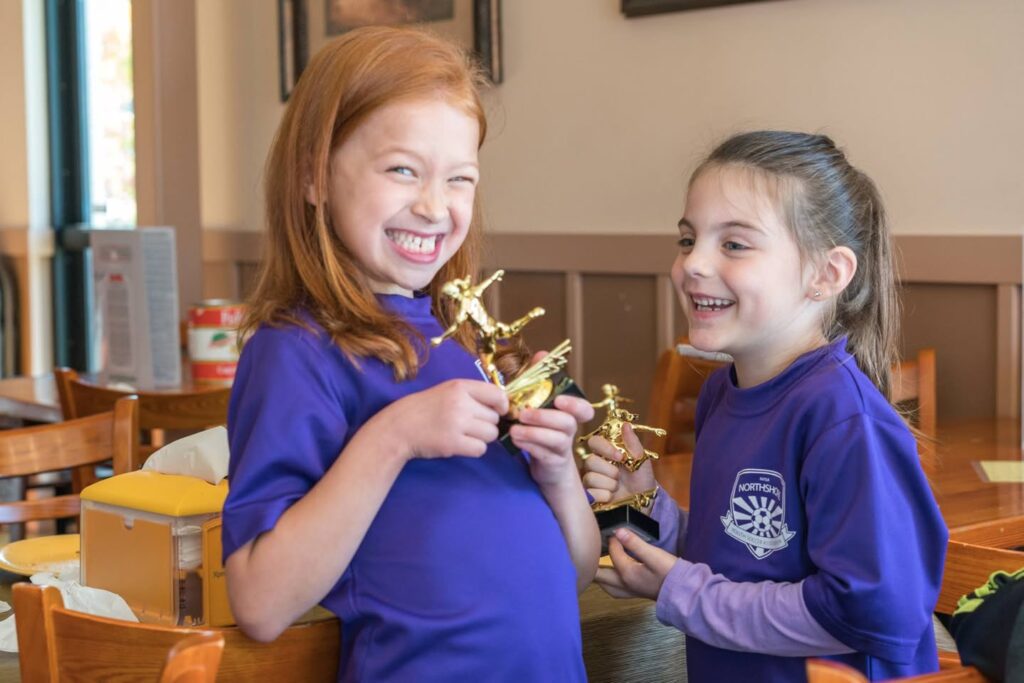 Two children wearing matching purple shirts smile while holding small golden figurines.