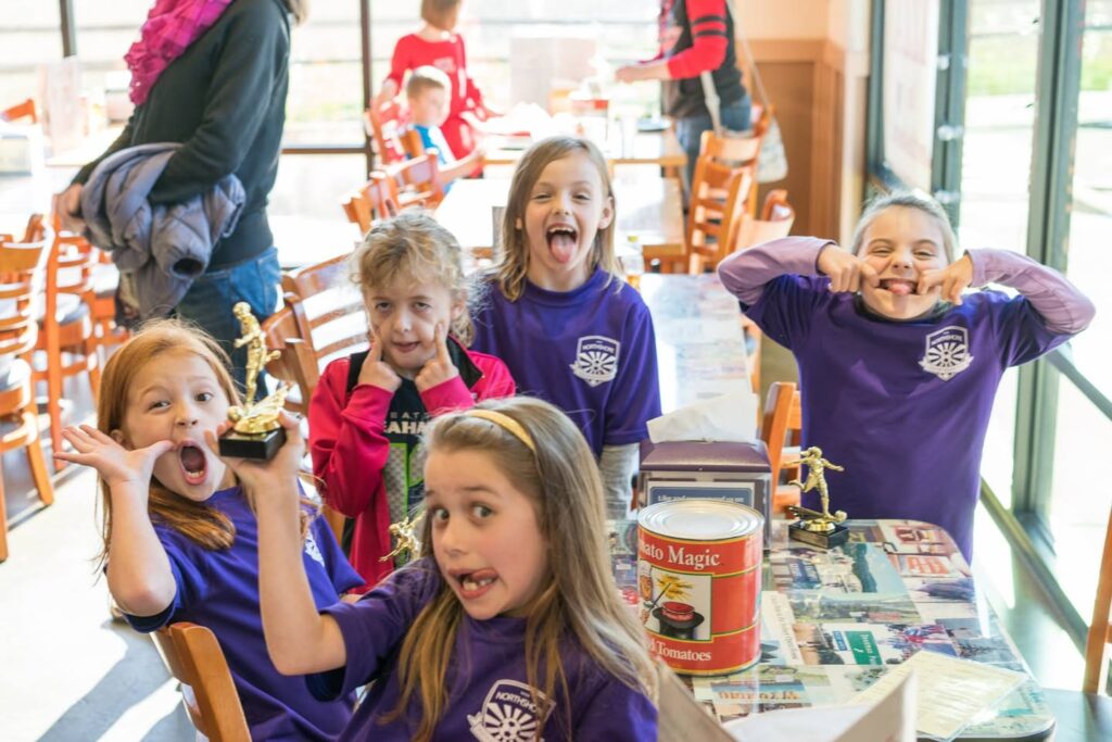 Children wearing purple shirts playfully cover their ears around a table full of snacks.