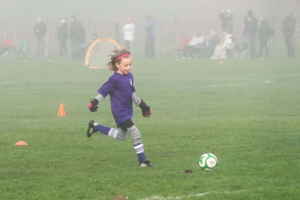 A young player in purple chases a soccer ball on a foggy field. Background spectators and orange cones are partially visible through the mist.