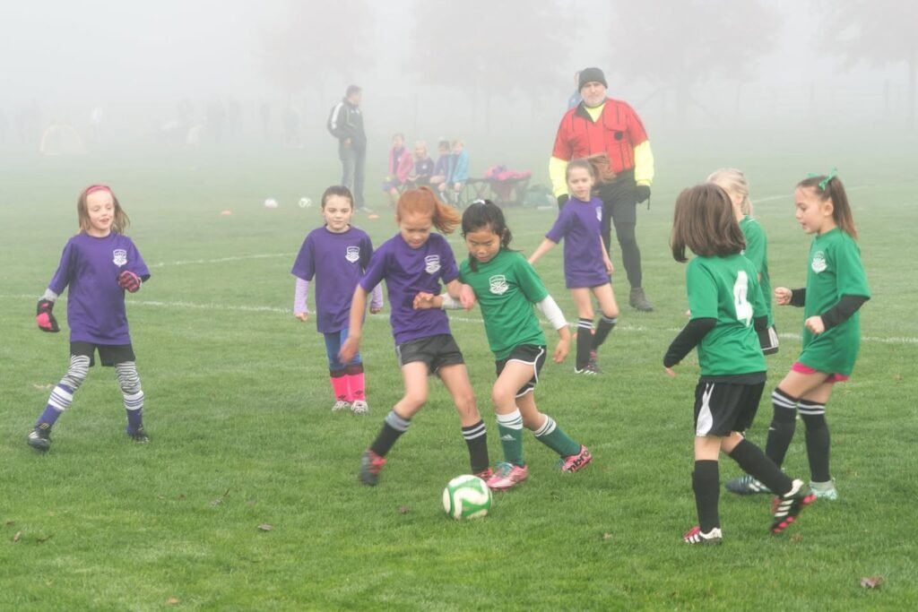 Two teams of children in purple and green uniforms play soccer on a grassy field in misty weather.