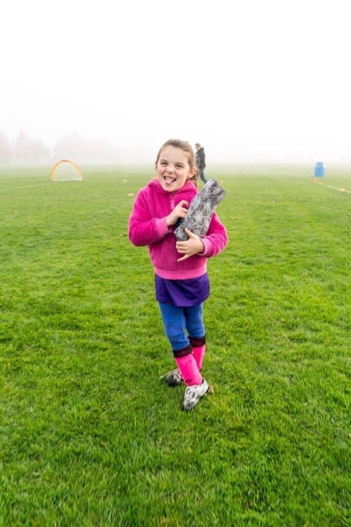A joyfilled child in a pink top and blue pants holding a large stick in a foggy grassy field. Warm outdoor activity in the misty morning light.