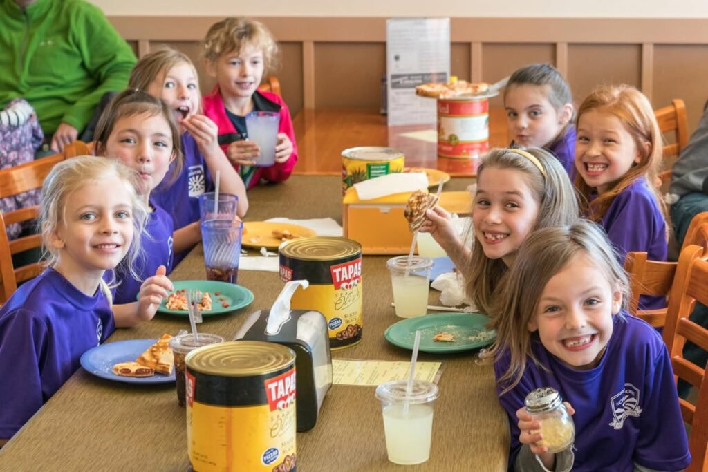 A group of children in purple shirts enjoy snacks and a fish feeding activity at a table.