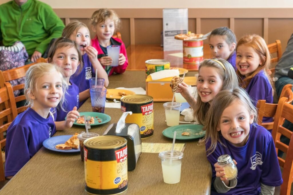 A group of children sit at a table sharing a meal of pizza and drinks in a casual cafeteria setting.