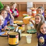 A group of children sit at a table sharing a meal of pizza and drinks in a casual cafeteria setting.