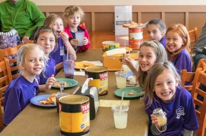 A group of children sit at a table sharing a meal of pizza and drinks in a casual cafeteria setting.