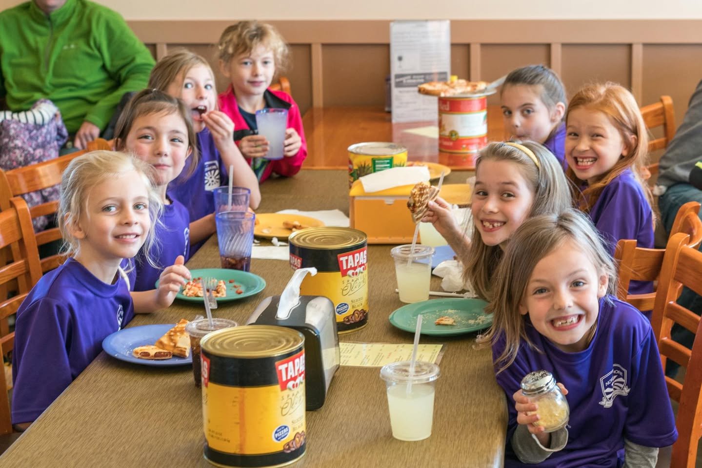 A group of children sit at a table sharing a meal of pizza and drinks in a casual cafeteria setting.