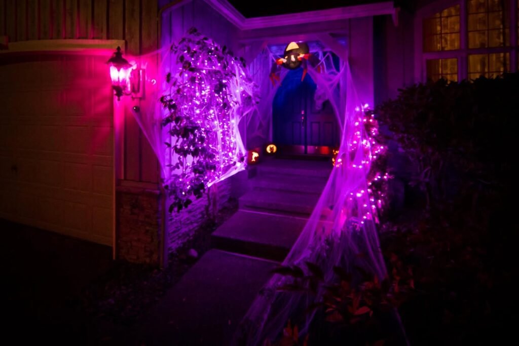 A creepy nighttime view of a house entrance illuminated by vibrant purple and pink lights, highlighting a glowing bush and a lantern.