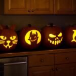 Four carved pumpkins with cat, bat, zombie, and dolphin designs sit on a wooden kitchen counter.
