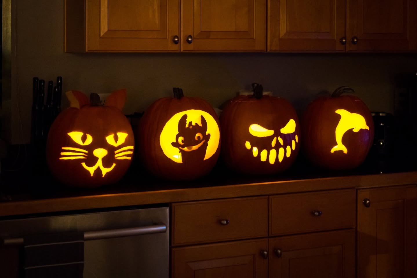 Jack O’Lanterns Four carved pumpkins with cat, bat, zombie, and dolphin designs sit on a wooden kitchen counter.