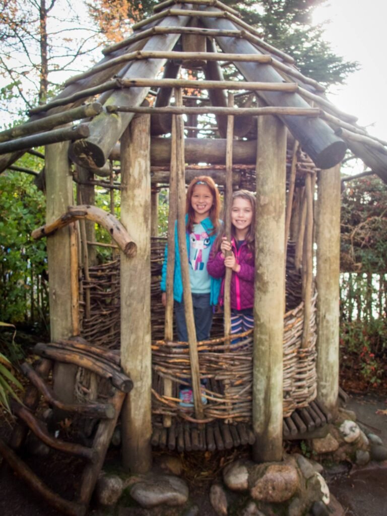 Two children smiling from inside a rustic treehouse structure built from logs and branches.