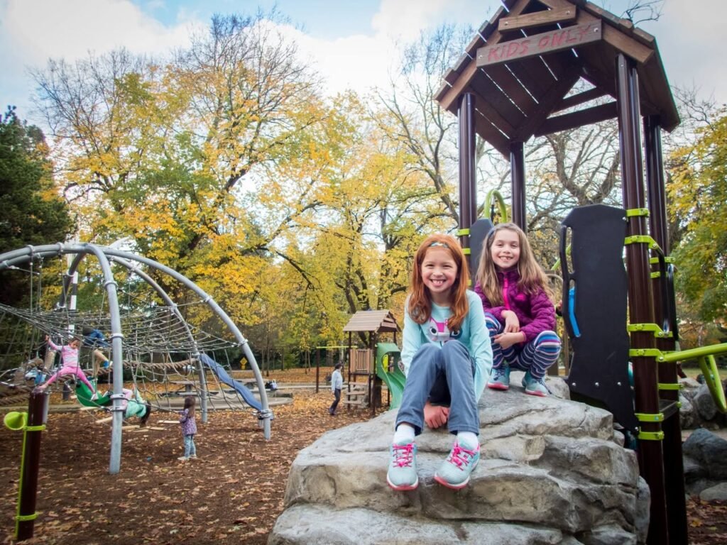 Two children sit together on stone steps at a playground surrounded by autumn trees and colorful equipment.