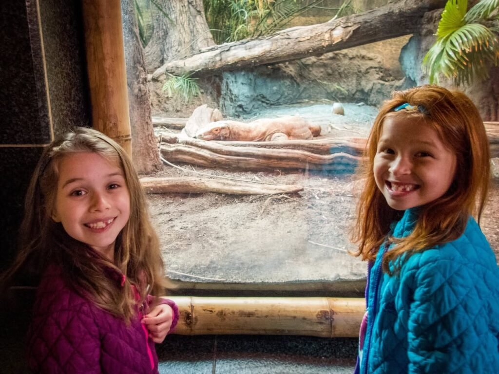 Two smiling children standing by a zoo enclosure with a white bear resting in the background.