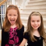 Two young girls smile and laugh on a carpeted staircase inside their home. The setting is a bright, white stairwell in a family home.