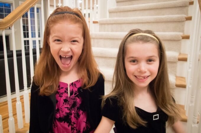 Two young girls smile and laugh on a carpeted staircase inside their home. The setting is a bright, white stairwell in a family home.