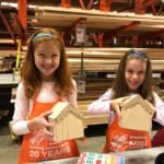 Two children wearing red aprons proudly display their handmade birdhouses at a Home Depot Kids Workshop.