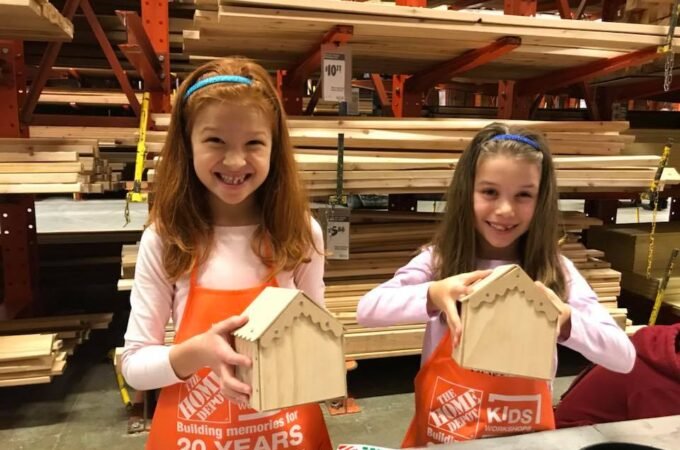 Two children wearing red aprons proudly display their handmade birdhouses at a Home Depot Kids Workshop.