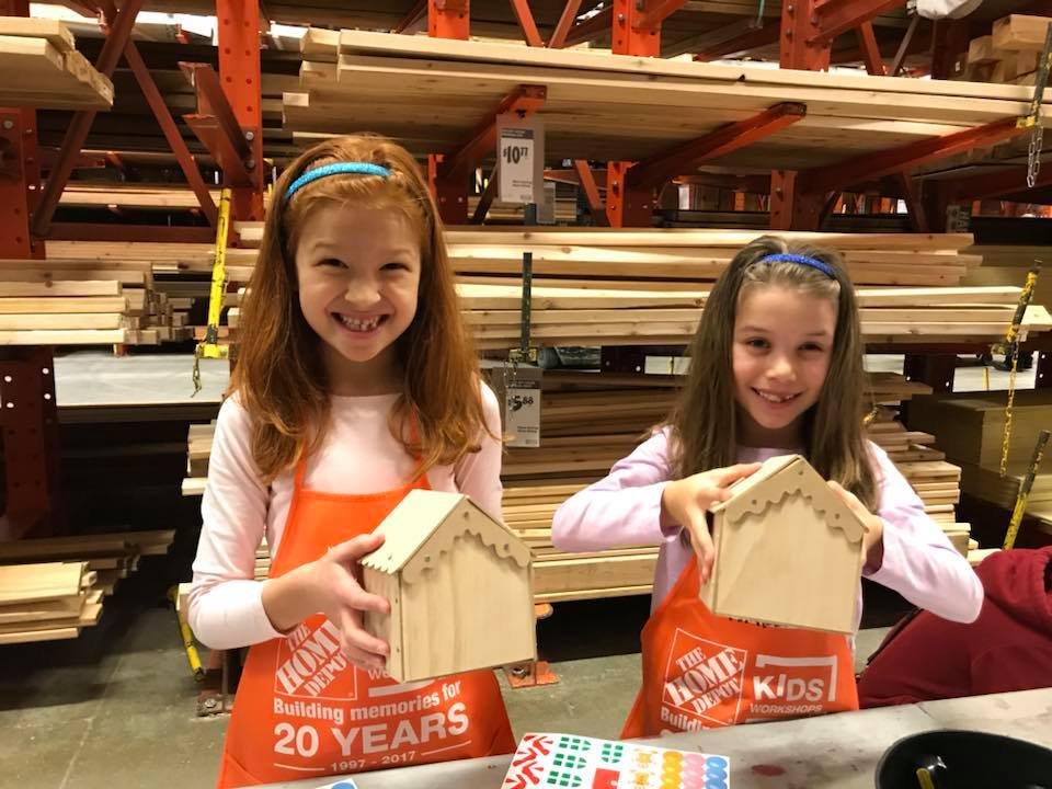 Two children wearing red aprons proudly display their handmade birdhouses at a Home Depot Kids Workshop.