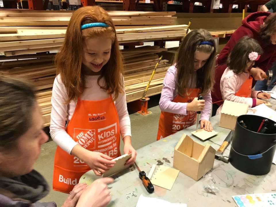 Children wearing orange aprons assemble small wooden boxes at a workbench in a hardware store.
