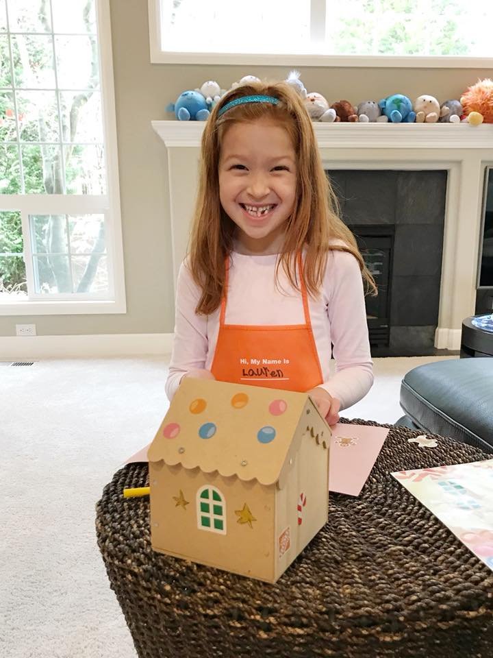 A smiling individual wearing an orange apron displays a handmade cardboard gingerbread house near a fireplace.