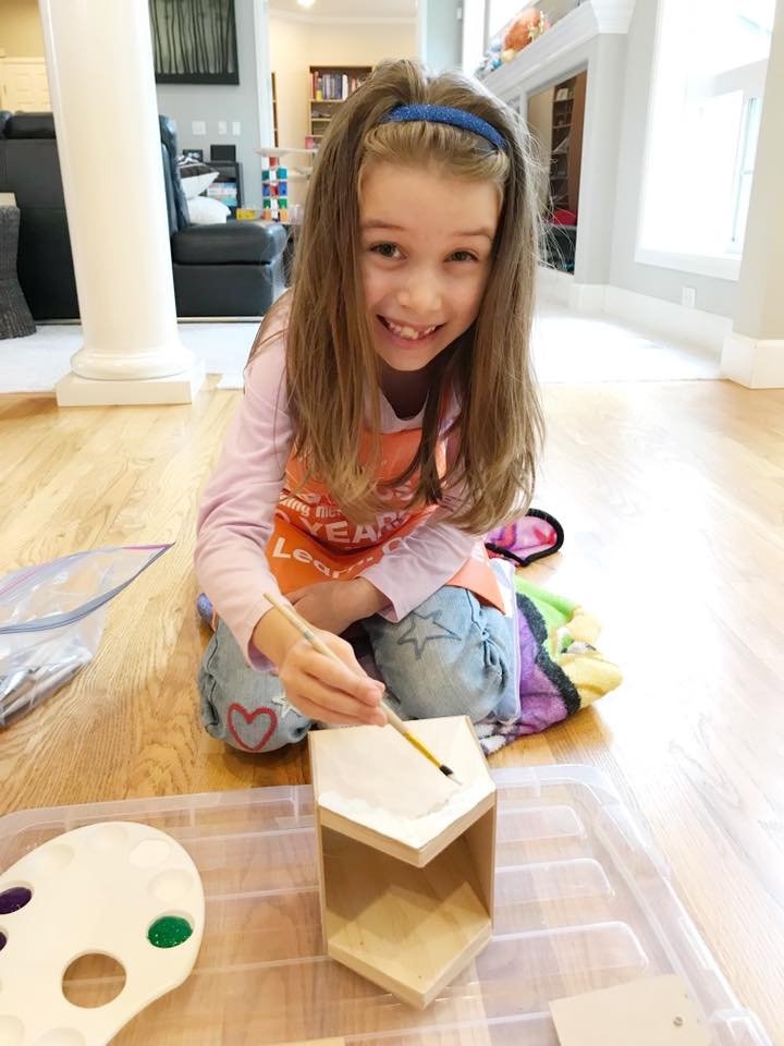 A young girl paints a small wooden block on a protective sheet in a bright living room with hardwood floors.