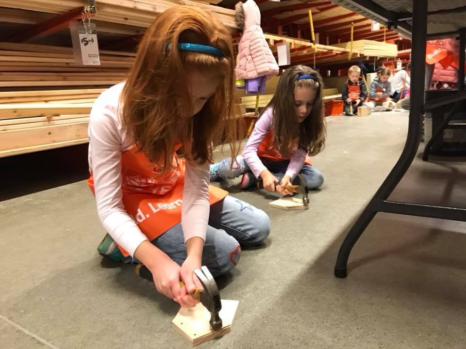 Two children in orange aprons practicing sanding on a floor panel at a hardware store. They are wearing safety headbands, focused on their task with tools.