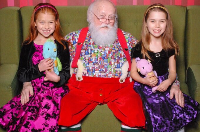 Elderly man dressed as Santa Claus sits with two young girls holding stuffed animals on a green couch with a holiday wreath in the background.
