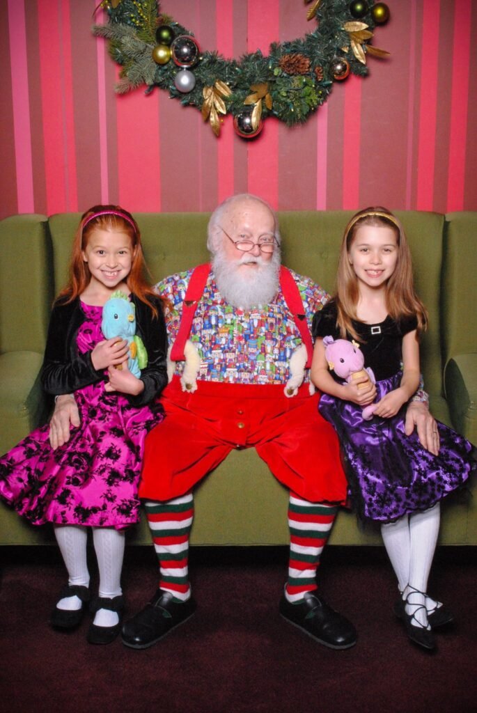 Elderly man dressed as Santa Claus sits with two young girls holding stuffed animals on a green couch with a holiday wreath in the background.