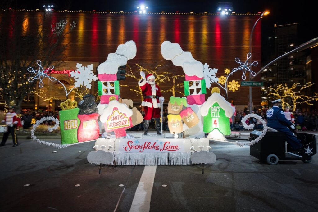 A colorful Christmas parade float featuring Santa Claus and festive houses, captured at night with string lights and snowflake decorations in the Pacific Northwest.
