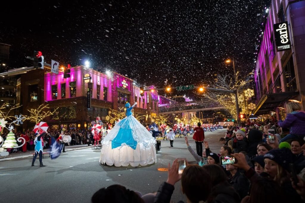 A festive evening street parade features an illuminated float in a snowy setting. Spectators line the street capturing photos of the vibrant event.