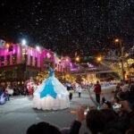 A festive evening street parade features an illuminated float in a snowy setting. Spectators line the street capturing photos of the vibrant event.