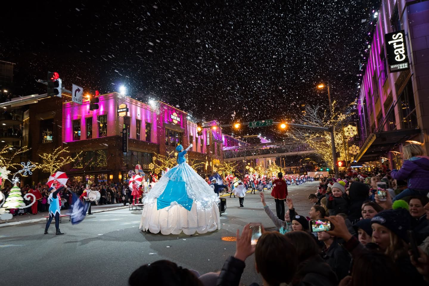 A festive evening street parade features an illuminated float in a snowy setting. Spectators line the street capturing photos of the vibrant event.