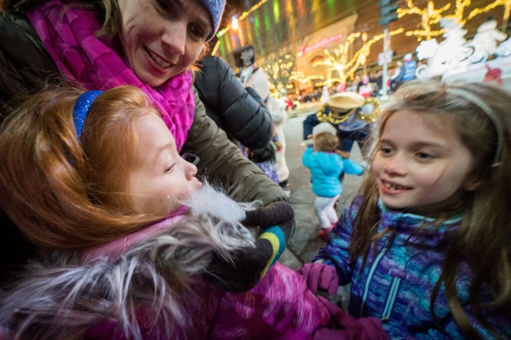 A family enjoys a festive winter evening with bright holiday light displays in the background. Children wearing colorful jackets and scarves explore the illuminated event.