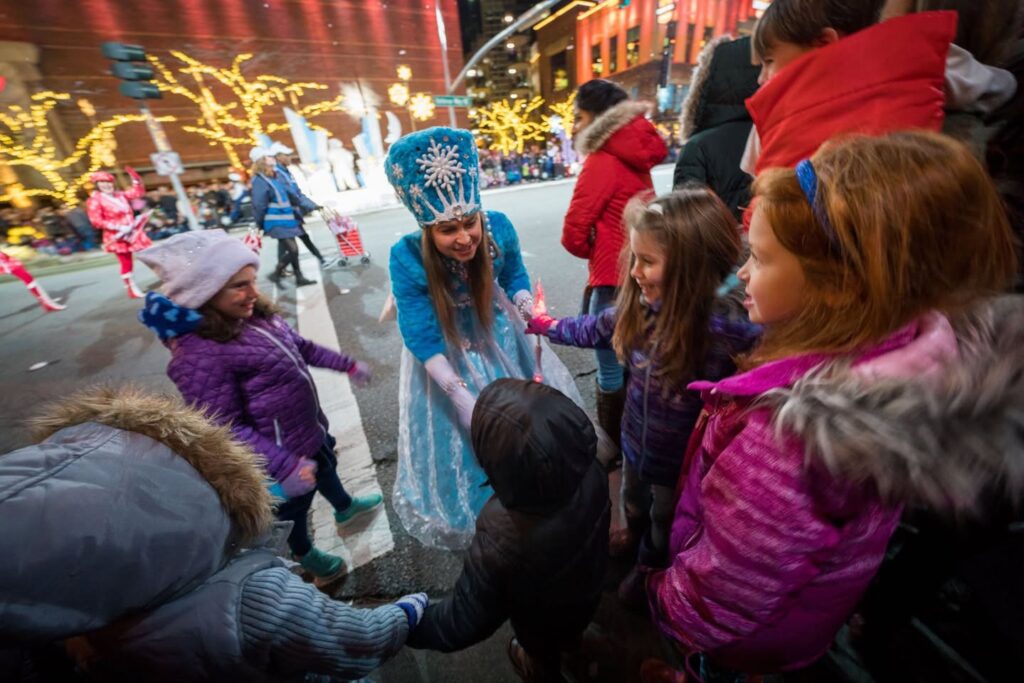 A costumed individual interacts with children during a festive winter event with holiday lights. Snow Queen offers handshakes to excited young attendees outdoors.
