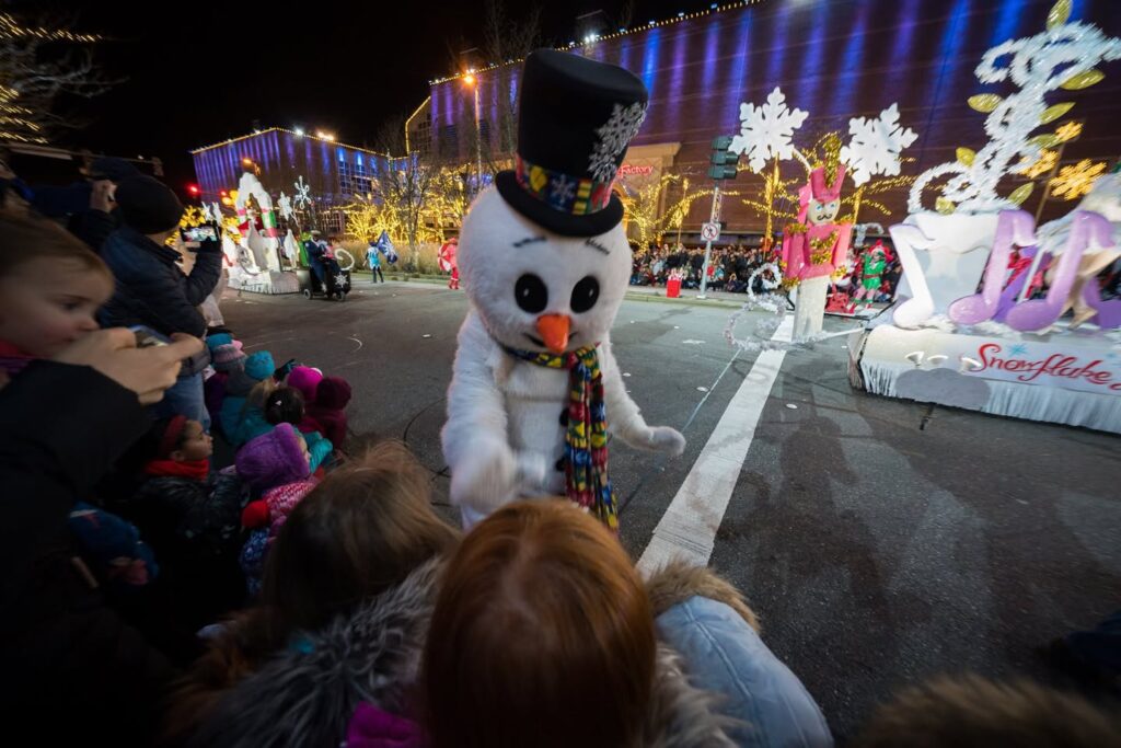 A festive snowman character wearing a top hat and scarf walks along a parade float decorated with lights.