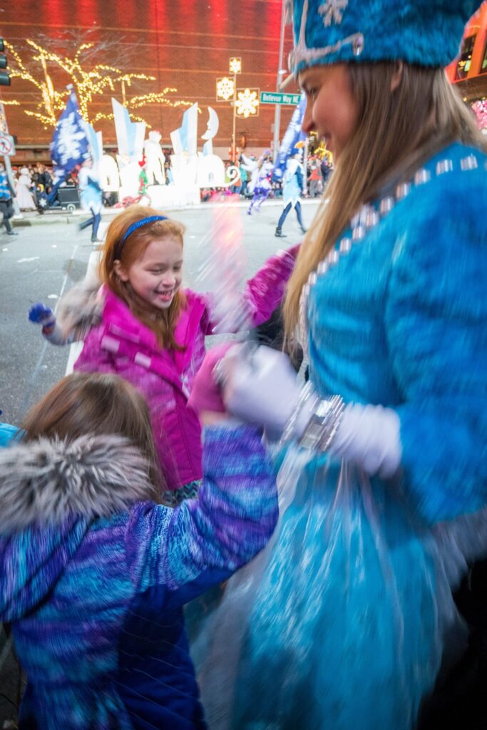 An adult in a vibrant blue winter costume interacting joyfully with children in colorful coats at a festive winter event.