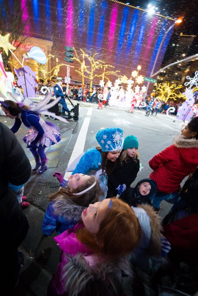 A crowd of people bundled in winter gear admires a colorful light display during a nighttime holiday parade.