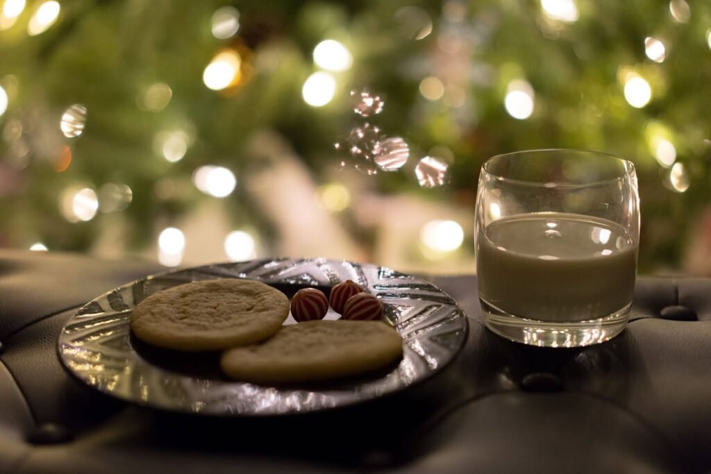 A plate of gingerbread cookies and peppermints sits beside a glass of eggnog in front of a lit Christmas tree.