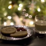 A plate of gingerbread cookies and peppermints sits beside a glass of eggnog in front of a lit Christmas tree.