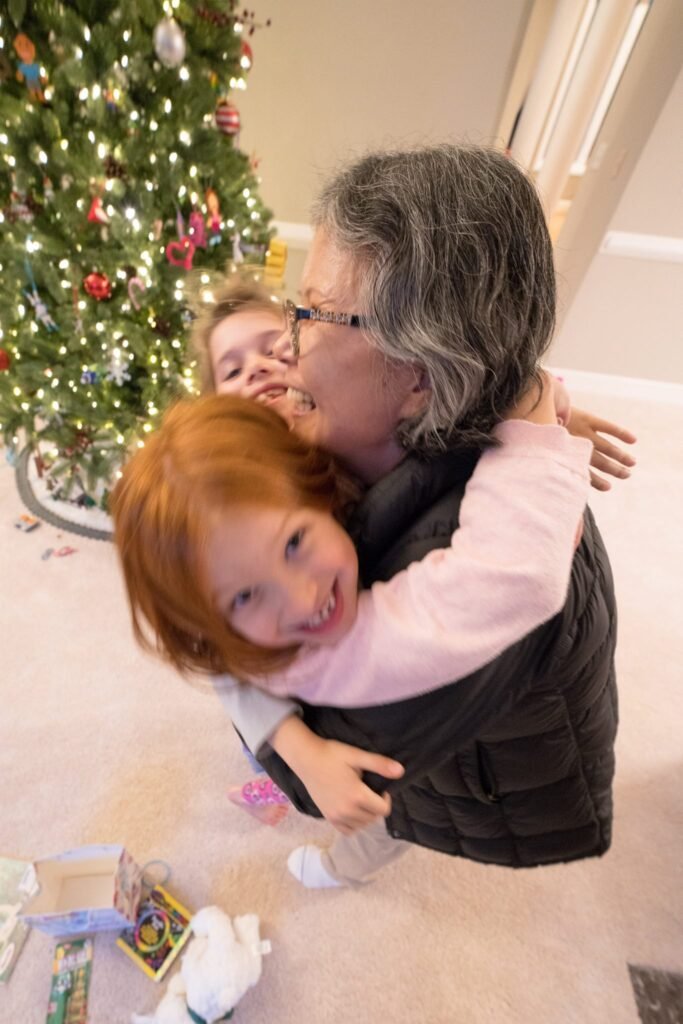 A woman and young child share a warm hug by a decorated Christmas tree filled with colorful lights and ornaments in a cozy home setting.