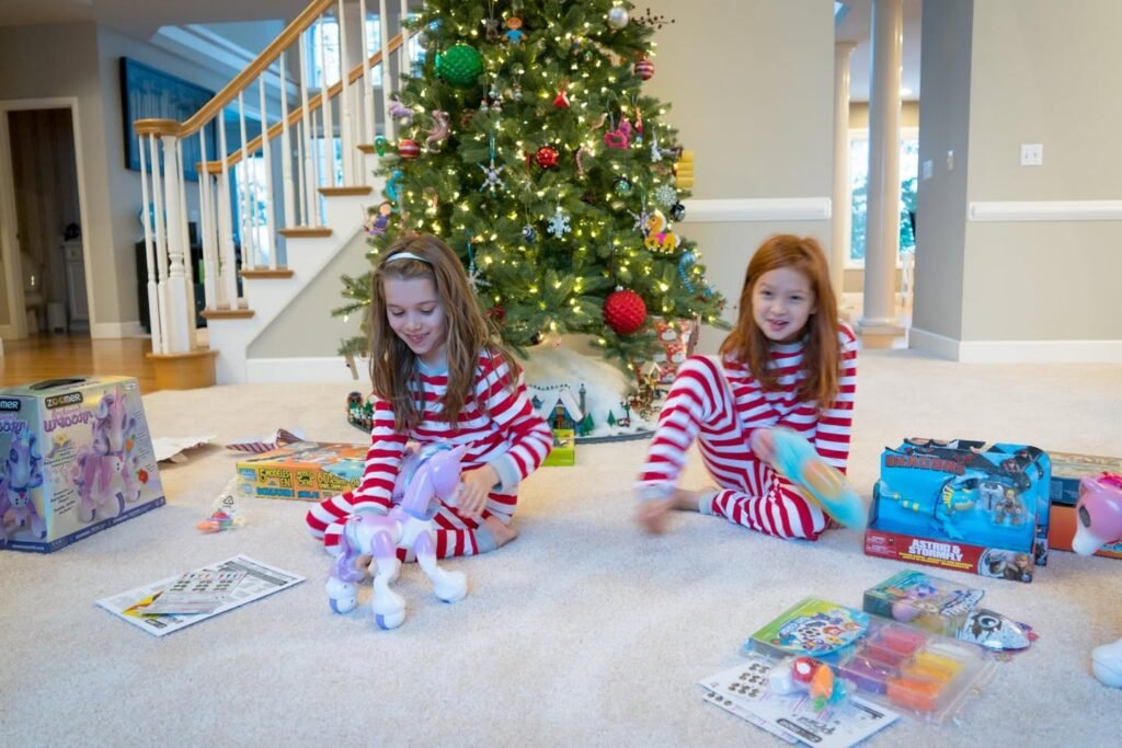 Two children in matching red and white striped pajamas play with toys on a carpet below a decorated Christmas tree. A grand staircase and bright living area are visible in the background.