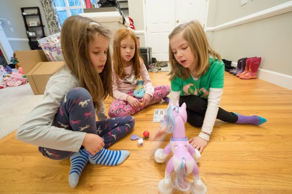 Three children sit on the wooden floor playing a board game with a pink toy unicorn. They are focused on their board game during a cozy evening at home.