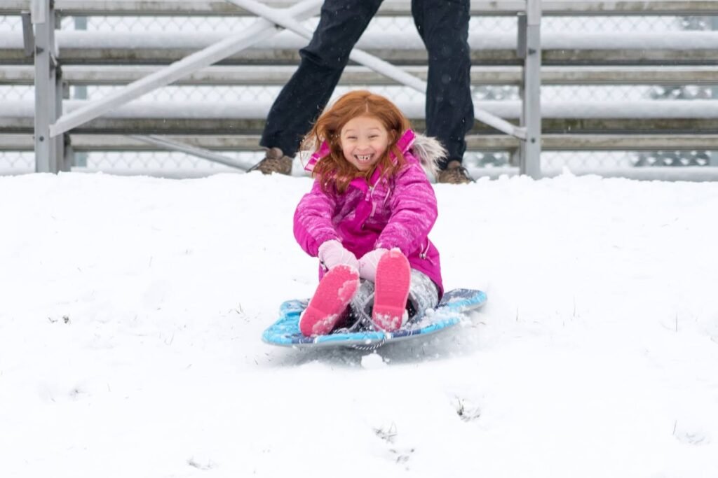 A child in a pink jacket slides down a snowy hill on a blue sled.