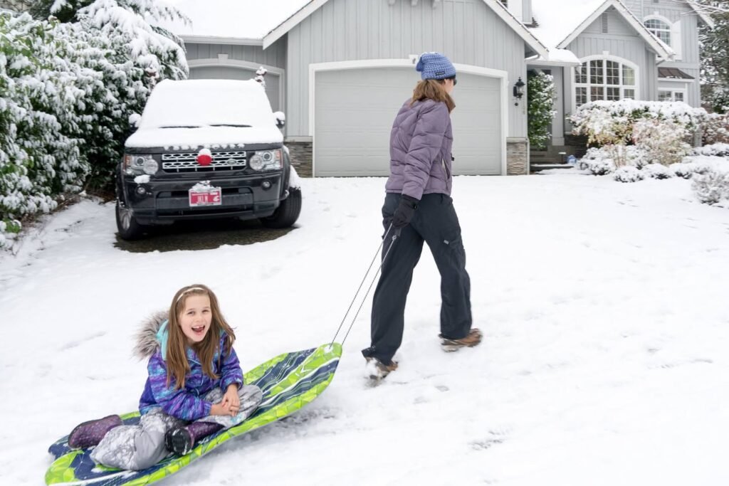 A child rides a bright sled through snow on a driveway while an adult walks nearby in a winter jacket. The scene features a snow-covered house, bushes, and a parked SUV.