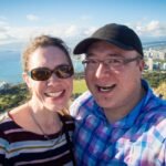 Two travelers taking a selfie at a scenic overlook, with the coastline and an urban cityscape visible in the background.